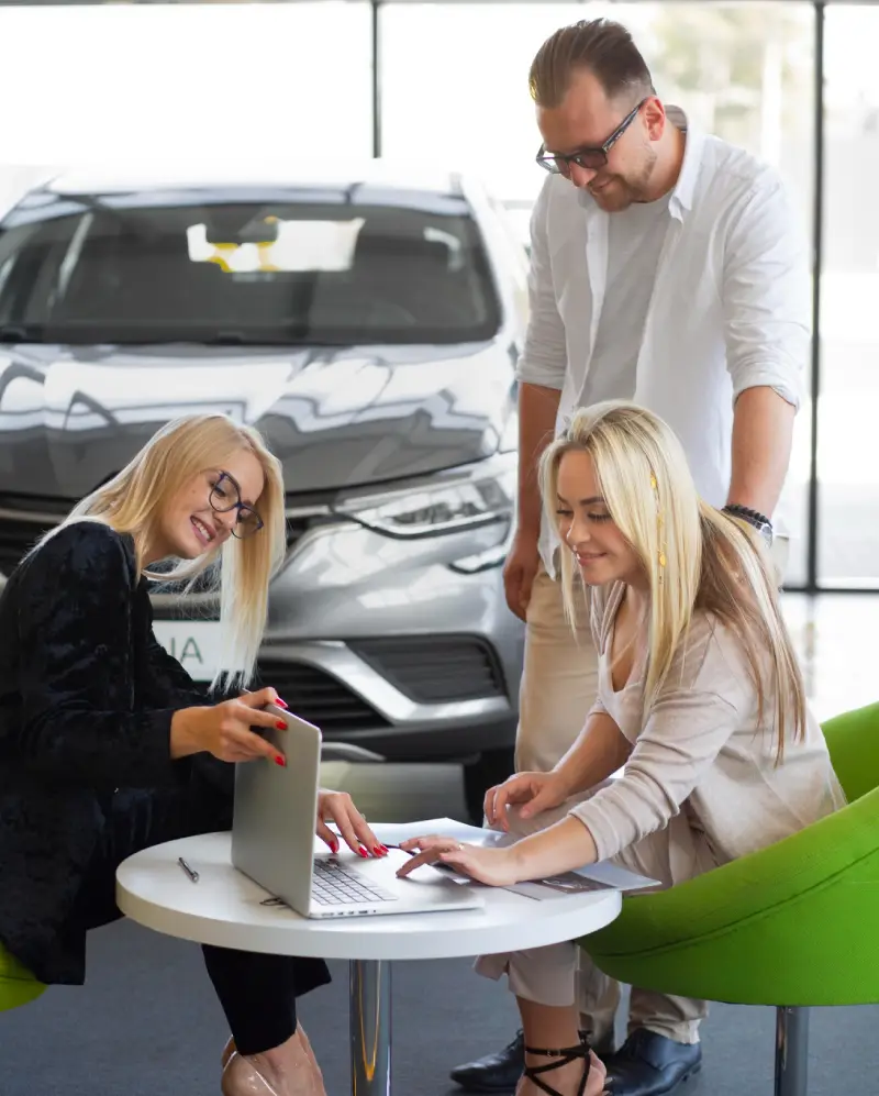 img-7-1 - new Three individuals collaborate at a table with a laptop in a car dealership, while a sleek vehicle is prominently displayed in the background.