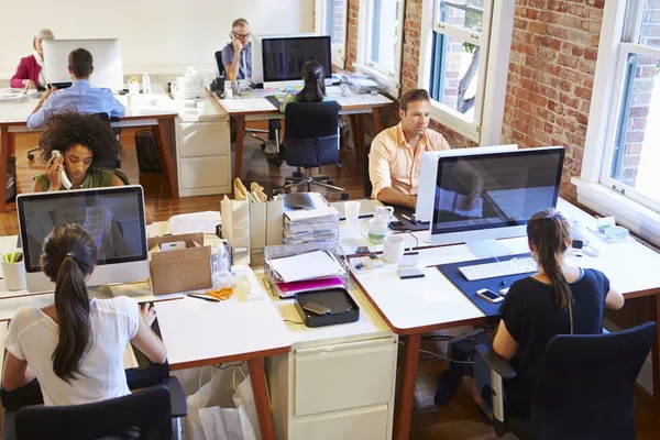 A modern office setting with several employees working at desks, using computers, surrounded by large windows and exposed brick walls.