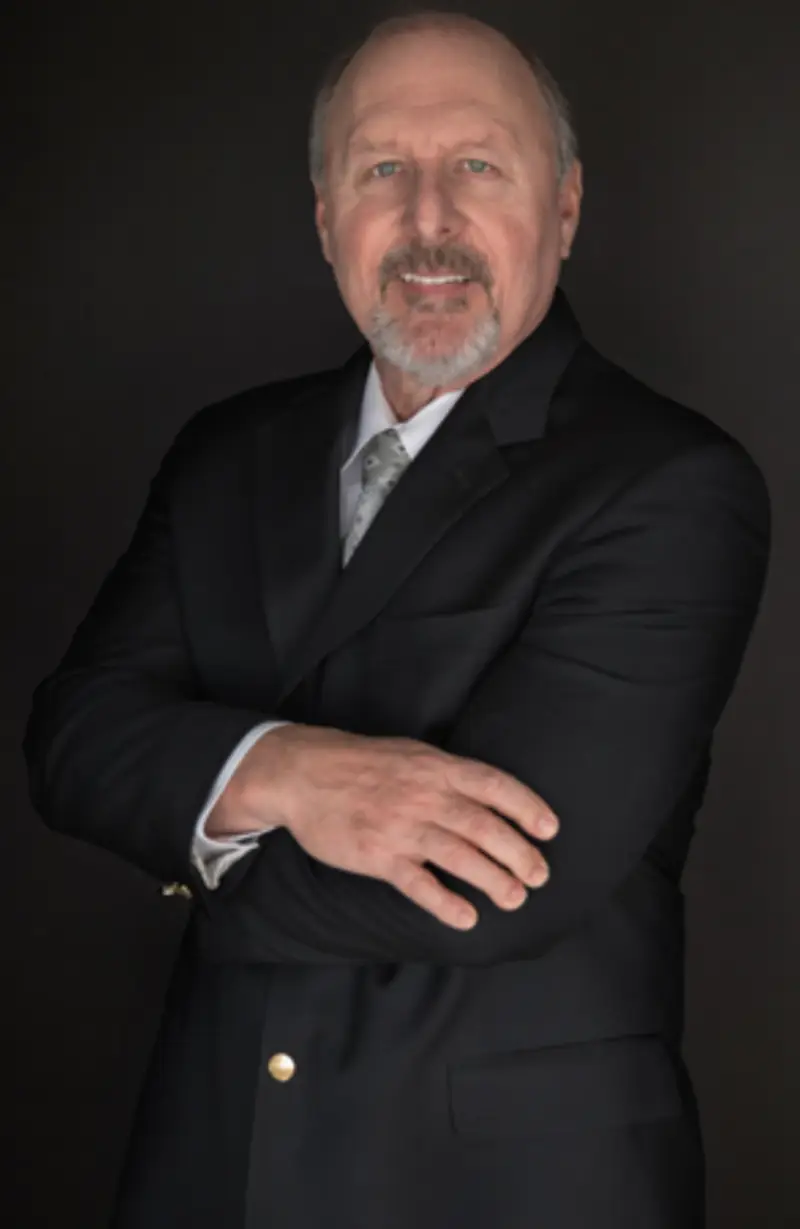 profile pic A man in a formal black suit stands confidently with his arms crossed, set against a dark background.